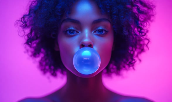 Young woman blowing bubble gum in vibrant lighting. A young woman with curly hair is playfully blowing a bubble with blue gum, illuminated by pink and purple lights. photo