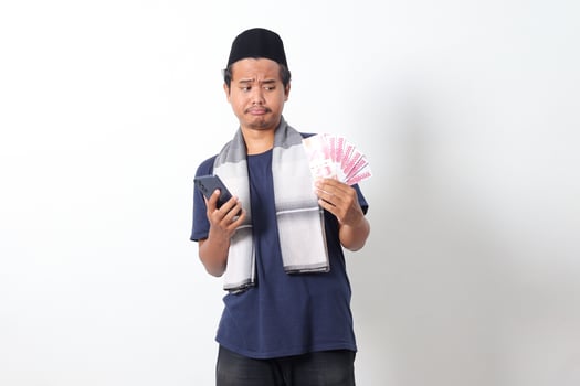 Portrait of shocked Asian muslim man wearing sarung scarf holding rupiah money banknote while holding and looking at his mobile phone. Isolated image on white background photo