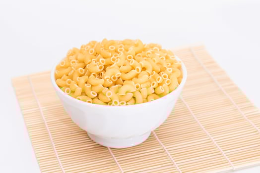 Uncooked Chifferi Rigati Pasta in White Cup on Bamboo Mat on White Background. Fat and Unhealthy Food. Scattered Classic Dry Macaroni. Italian Culture and Cuisine. Raw Pasta photo
