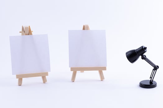 Two Wooden Easels Miniature with Blank White Square Canvas for Artists and Painters - Mockup. Mini Wooden Stands with Clean Artboard and Small Black Table Lamp on White Background, Copy Space photo