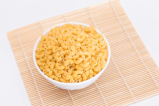 Uncooked Chifferi Rigati Pasta on White Cup on Bamboo Mat on White Background. Fat and Unhealthy Food. Scattered Classic Dry Macaroni. Italian Culture and Cuisine. Raw Pasta photo