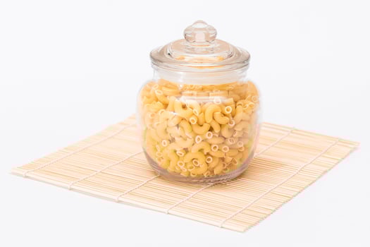 Uncooked Chifferi Rigati Pasta in Glass Jar on Bamboo Mat Isolated on White Background. Fat and Unhealthy Food. Scattered Classic Dry Macaroni. Italian Culture and Cuisine. Raw Pasta - Isolation photo