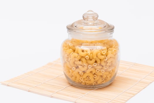 Uncooked Chifferi Rigati Pasta in Glass Jar on Bamboo Mat on White Background. Fat and Unhealthy Food. Classic Dry Macaroni. Italian Culture and Cuisine. Raw Pasta photo