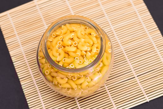 Uncooked Chifferi Rigati Pasta in Glass Jar on Bamboo Mat on Black Background. Fat and Unhealthy Food. Classic Dry Macaroni. Italian Culture and Cuisine. Raw Pasta photo