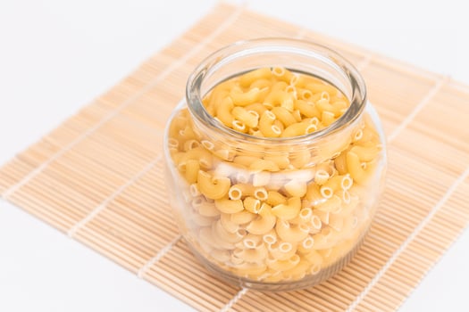 Uncooked Chifferi Rigati Pasta in Glass Jar on Bamboo Mat on White Background. Fat and Unhealthy Food. Classic Dry Macaroni. Italian Culture and Cuisine. Raw Pasta photo