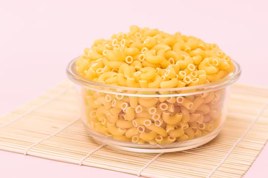 Uncooked Chifferi Rigati Pasta in Glass Jar on Bamboo Mat on Pink Background. Fat and Unhealthy Food. Classic Dry Macaroni. Italian Culture and Cuisine. Raw Pasta photo