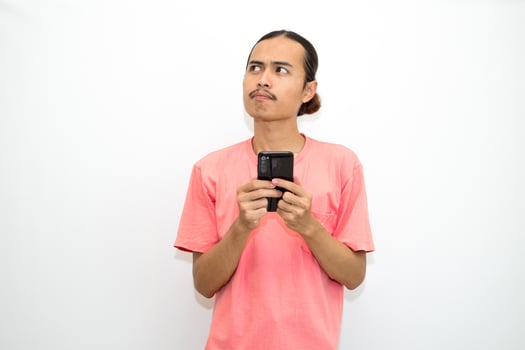 Asian, Indonesian man standing holding cellphone with hand on chin as if thinking hard, thinking about work or school assignment to find solution. Isolated on white background photo