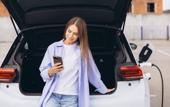 Woman Leaning Against Electric Car Holding Smartphone In Urban Parking Lot photo