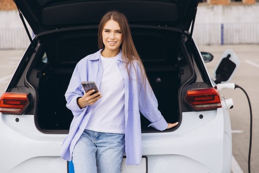 Woman Leaning Against Electric Car Holding Smartphone In Urban Parking Lot photo