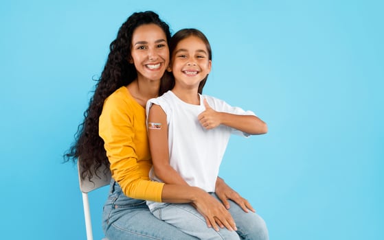 Arabic Girl And Her Mom Showing Vaccinated Arm And Thumbs Up Gesture After Covid-19 Vaccination Sitting Over Blue Background, Studio Shot. Coronavirus Protection For Kids Concept photo