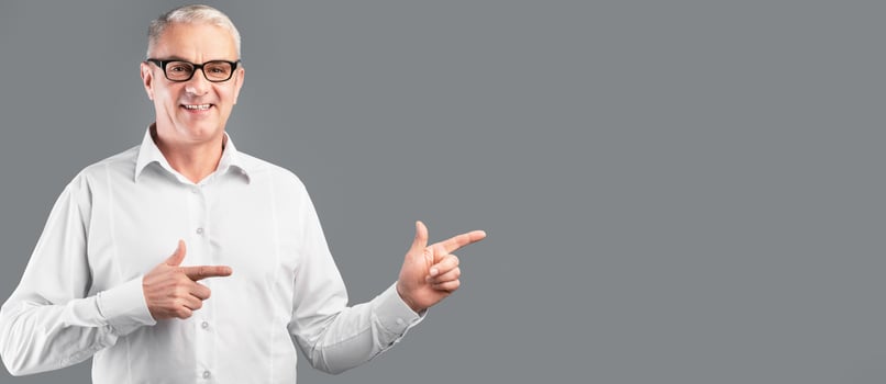 Cool Offer. Smiling mature man in glasses pointing two index fingers aside at copyspace, gray studio wall, panorama photo