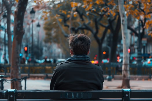 A man sits alone on a bench, gazing out at a vibrant autumn park filled with trees. The city lights begin to glow as dusk approaches, creating a calm atmosphere photo