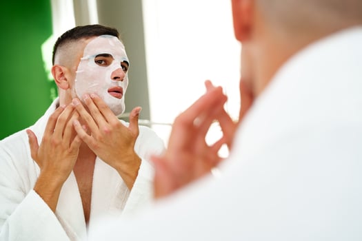 Handsome young man applying sheet mask near mirror in bathroom photo