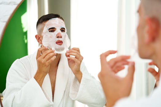 Handsome young man applying sheet mask near mirror in bathroom photo