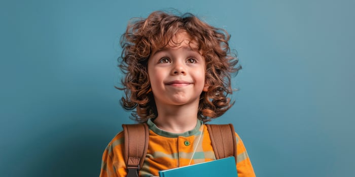 Young child with curly hair holding a book wears a backpack against a blue background photo