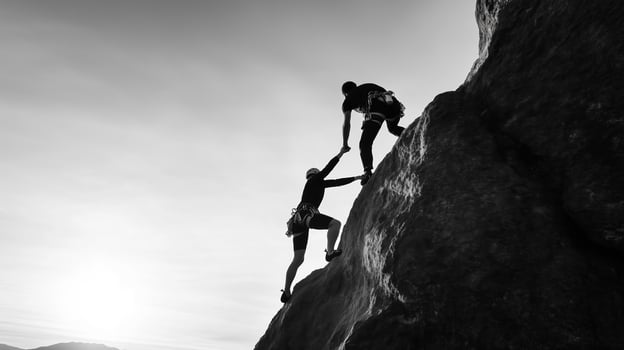 Two climbers ascending a rocky cliff during sunset, showcasing teamwork and determination in a dramatic black and white landscape photo