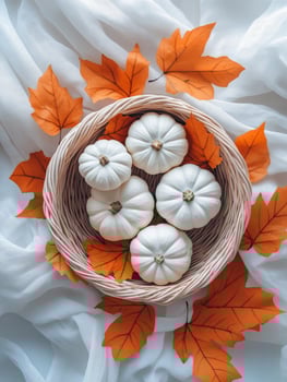 White pumpkins arranged in a wicker basket with autumn leaves on a delicate fabric backdrop photo