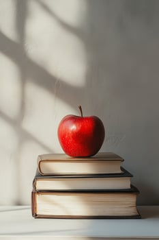 A vibrant red apple resting atop three stacked books on a sunny surface with shadows photo