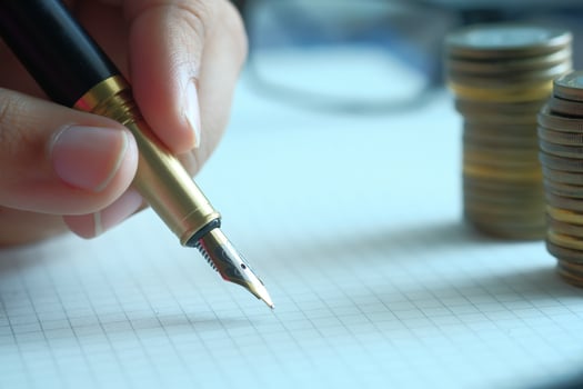 A closeup of a hand holding a fountain pen above a notepad with stacks of coins behind photo