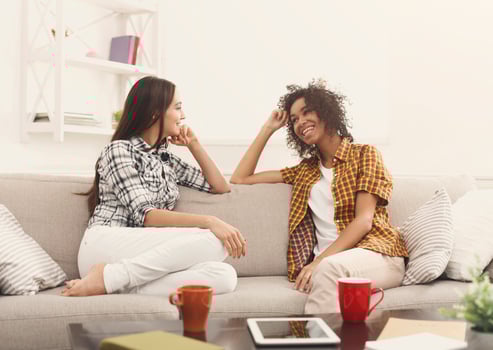 Two happy young female friends with coffee cups conversing in living room at home, chatting about their life and relations, gossip and slumber party concept, copy space photo