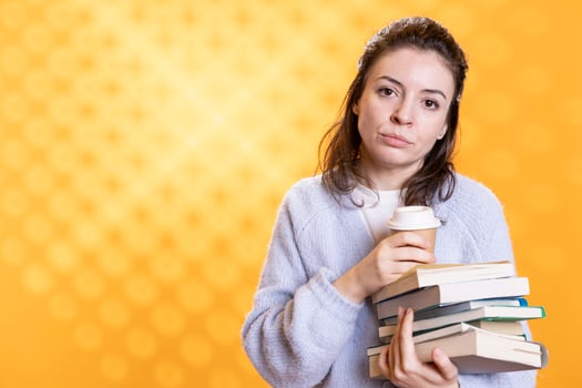 Portrait of tired woman with stack of books in hands and cup of coffee, isolated over studio background. Exhausted bookworm holding pile of novels and caffeine beverage, lacking energy photo