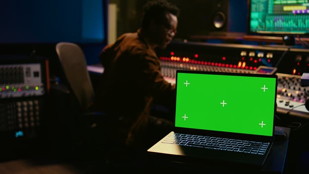 Skilled audio technician processing and mixing sounds next to mockup display on laptop, using variety on knobs, buttons and sliders to edit audio recordings and produce new music. Camera B. photo