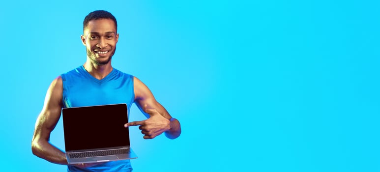 Online Gym Concept. Fit African Man Showing Laptop With Empty Screen For Your Text On Blue Studio Background. Mockup, Panorama photo