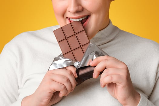 Positive Fatty Girl Biting Chocolate Bar, Enjoying Eating Sweets, Posing Over Yellow Background And Looking At Camera, Free Space photo