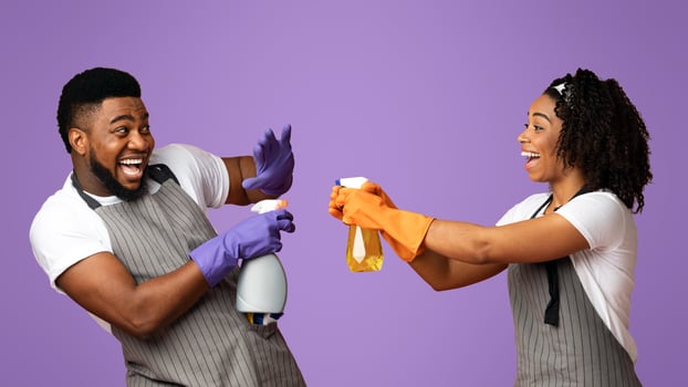 Cheerful Afro Couple In Aprons Playing With Spray Detergents, Playfully Aiming At Each Other While Posing On Pink Background In Studio photo
