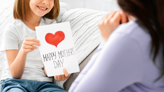Loving little daughter greeting her mother with Mother's Day, holding self made gift card, home interior photo