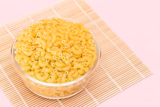 Uncooked Chifferi Rigati Pasta in Glass Jar on Bamboo Mat on Pink Background. Fat and Unhealthy Food. Classic Dry Macaroni. Italian Culture and Cuisine. Raw Pasta photo
