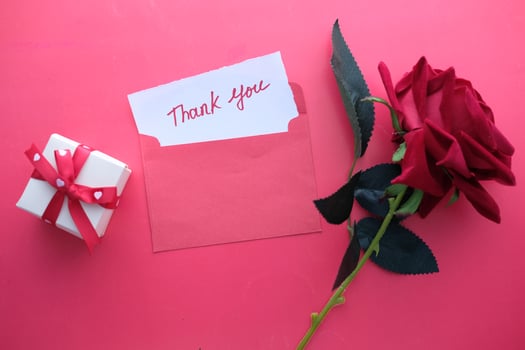 A Grateful Thank You Note Accompanied by a Red Rose and a Thoughtful Gift on a Pink Background photo