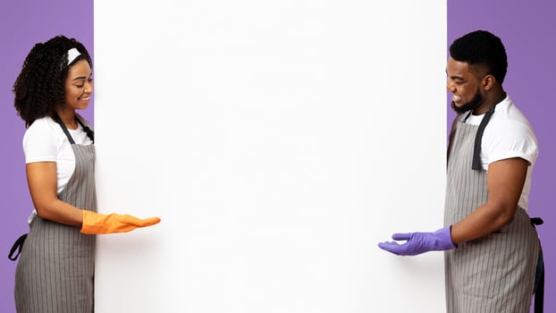 Spring Cleaning Tips. Positive Afro Couple In Aprons And Rubber Gloves Pointing At Big White Advertisement Board With Free Space For Text, Panorama photo