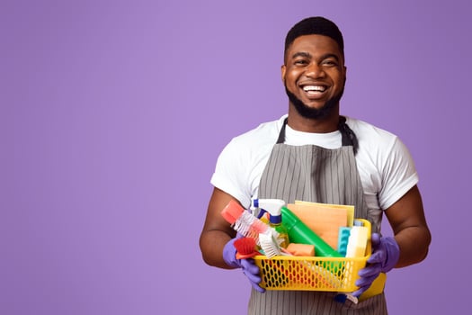 Portrait of positive black man holding basket with detergents and cleaning tools, isolated on pink background with copy space photo