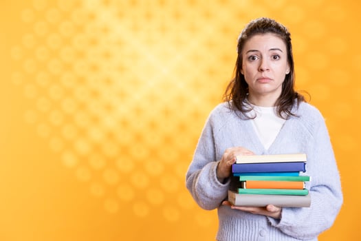 Portrait of woman with worried look on face carrying stack of books, gathering information for school exam, studio background. Student feeling anxious before university assessment, holding textbooks photo