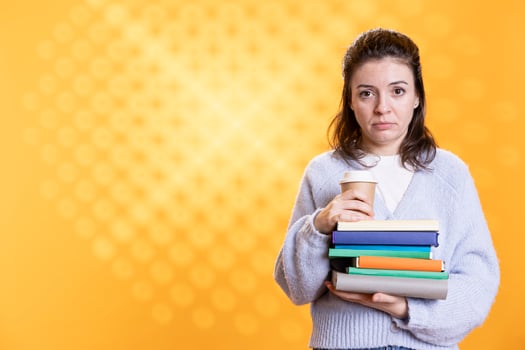 Portrait of woman with worried look on face carrying stack of books and coffee cup, studio background. Student feels anxious before university assessment, holding textbooks, drinking caffeine beverage photo