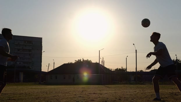 Silhouette of professional footballers kicking soccer ball with their heads on stadium at sunset. Two sportsmen showing tricks with ball while passing it to each other at field. Freestyle football photo