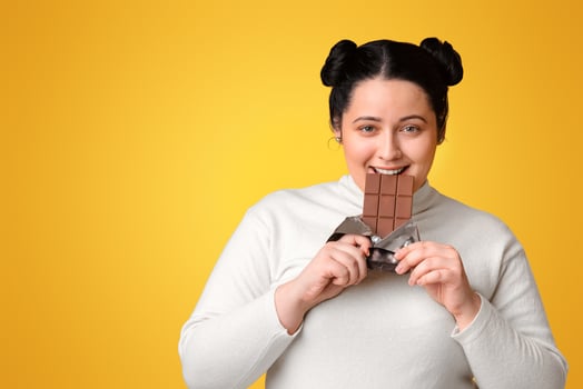 Pretty Plumb Girl Eating Milk Chocolate Bar Smiling To Camera Standing Over Yellow Studio Background, Empty Space photo