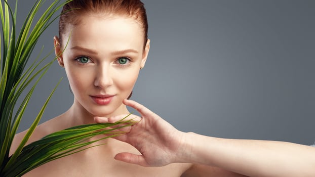 Ginger-Haired Young Lady Posing Touching Green Plant Looking At Camera Standing In Studio On Gray Background. Youth And Wellness. Natural Female Beauty Portrait Concept photo