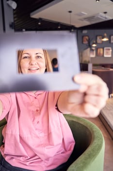 Smiling woman in pink shirt framing face with cardboard cutout in cozy cafe setting. photo