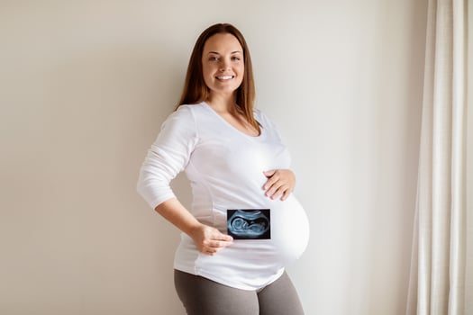 A smiling pregnant woman stands in a well-lit room, gently cradling her baby bump while holding an ultrasound picture in front of her belly, showcasing her excitement for motherhood. photo