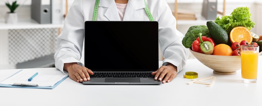 Happy african woman dietologist showing blank laptop screen, sitting at her cabinet, nutritionist consultation online photo