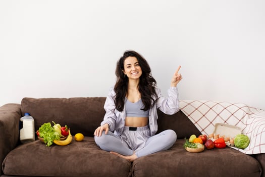 Young woman pointing up and showing healthy food on couch at home photo