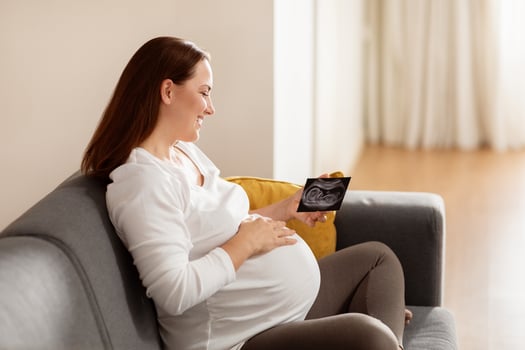A pregnant woman sits comfortably on a gray couch, smiling as she looks at an ultrasound photo. The soft afternoon light brightens the cozy living room around her. photo