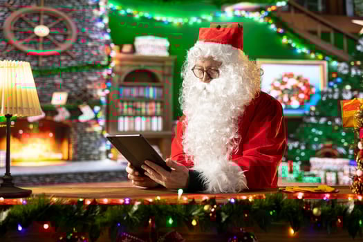 Santa Claus uses a tablet to navigate online webpages in his holiday decorated North Pole log cabin. Enjoying the festive and Christmas traditions, making gift delivery preparations. photo