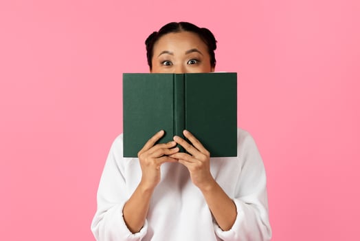 A woman with an excited expression playfully holds a green book in front of her face, against a vibrant pink background, showcasing a moment of joy and surprise. photo