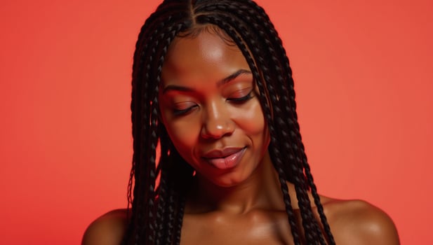 Closeup portrait of a radiant Black woman with long braids in a vibrant coral backdrop eyes closed gazing downwards photo