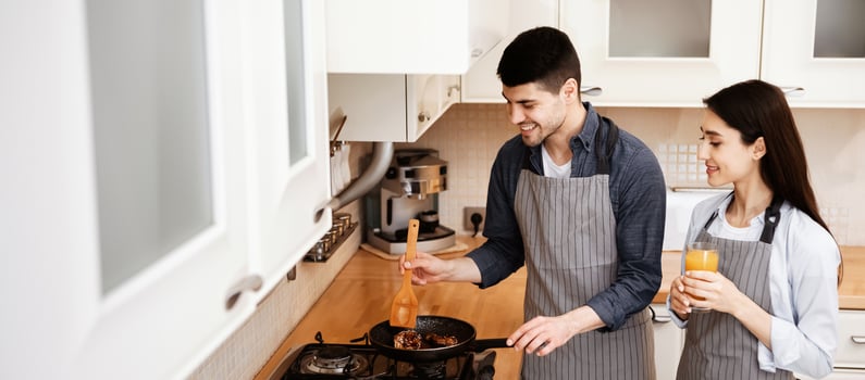 Cooking Romantic Dinner. Happy young man in apron frying meal on pan, girl holding glass with orange juice photo