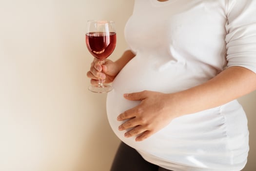 A pregnant individual stands indoors, gently cradling their belly with one hand while holding a glass of red wine in the other, surrounded by a serene atmosphere. photo