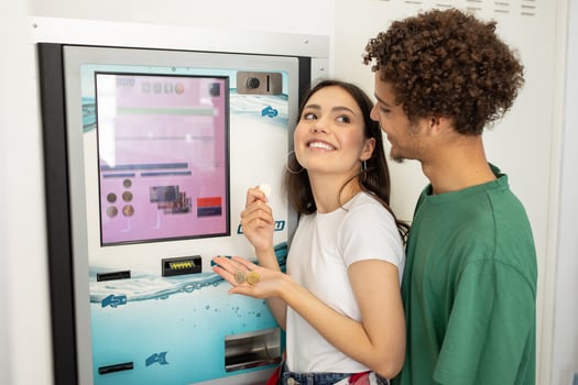 A smiling young woman holds snacks while her partner stands close, both enjoying a moment together at a vending machine in a well-lit area. photo